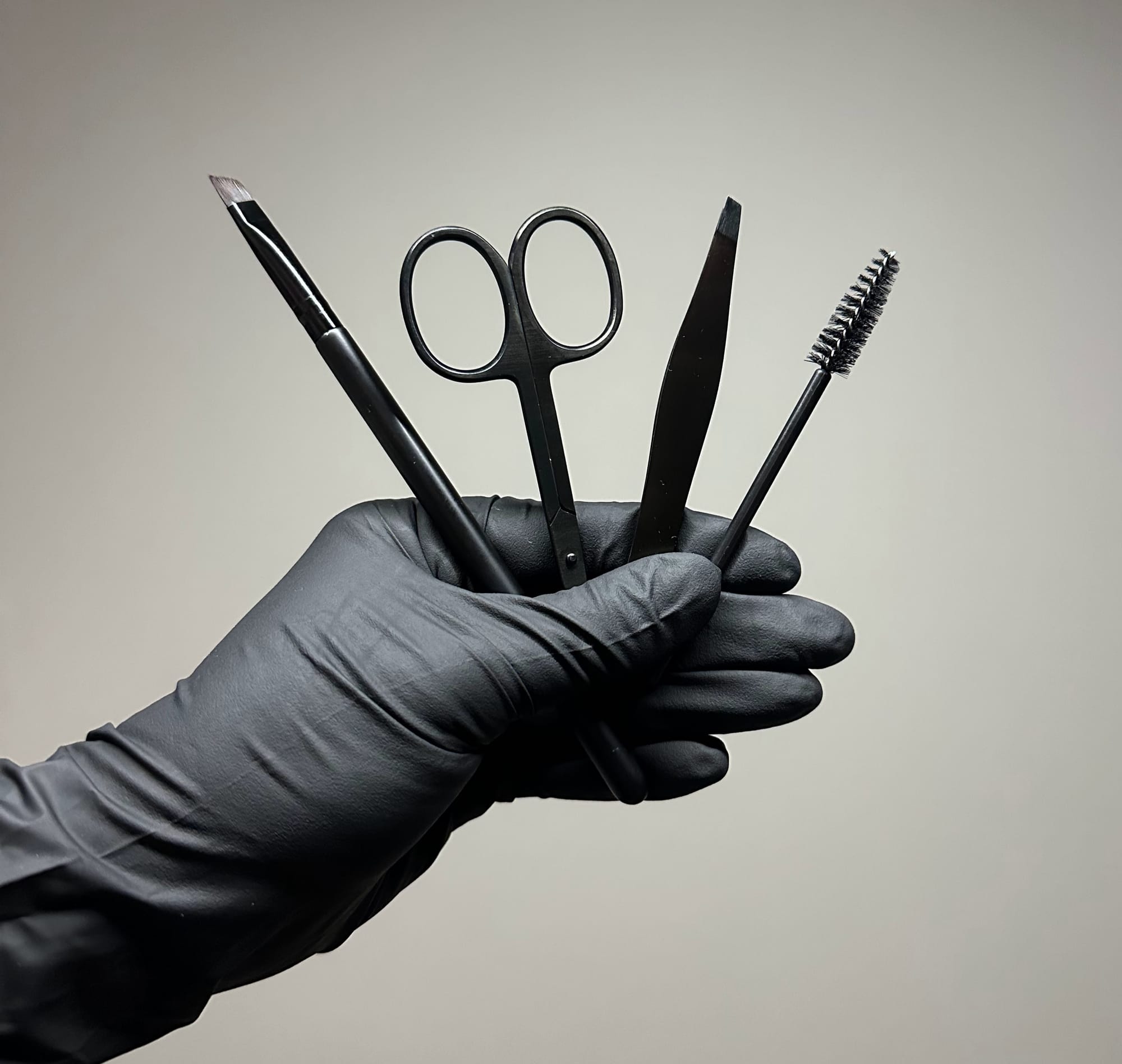 Wenkbrauwgereedschap in een gehandschoende hand: pincet, schaartje, borsteltje en potlood