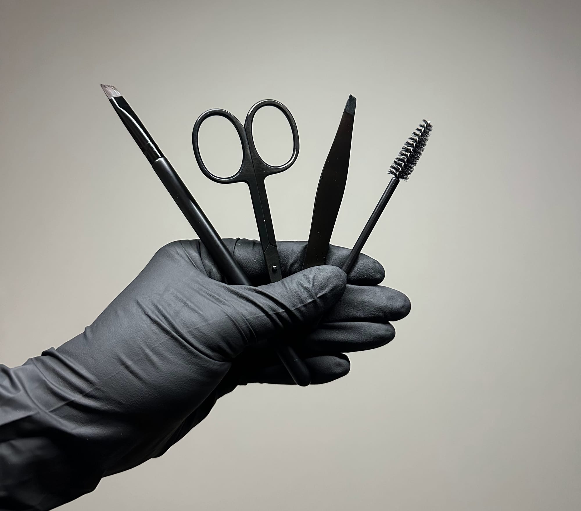 Wenkbrauwgereedschap in een gehandschoende hand: pincet, schaartje, borsteltje en potlood